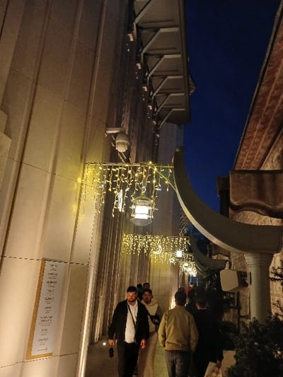 The image shows a narrow alleyway between two buildings in the Beyoğlu district of İstanbul, Türkiye. It is nighttime. String lights illuminate the alleyway. The building on the left is a modern, white building with a smooth, flat surface. The building on the right is older, with a rough, stone facade. The buildings are lined with strings of lights, and there are several people walking in the alleyway. The people are dressed casually, and some of them are carrying bags. The alleyway is empty except for the people. There is a sign on the wall of the building on the left that reads "Deniz". There is also a security camera mounted on the wall of the building on the left. The image is taken from the ground level, looking up towards the top of the alleyway.