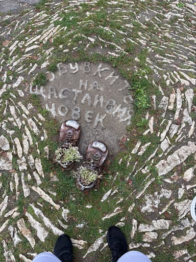 The image presents an overhead view of an outdoor ground installation in Lukovit, Bulgaria. A circular concrete plaque is centrally positioned, featuring raised Cyrillic lettering that translates to "SHOES OF A HAPPY PERSON". Resting on this plaque are two dark brown, shoe-shaped planters, appearing to be crafted from metal or a metallic-finished material, each containing a small, green flowering plant with white blossoms. The plaque is integrated into a rustic pathway composed of irregularly shaped, light-colored stones set into dirt, with patches of green grass growing between the stones and around the concrete. In the immediate foreground, the lower legs and feet of a person are visible, wearing black athletic shoes with white socks or pant cuffs, standing over the described ground feature.