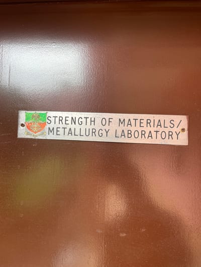 The image shows a metal sign affixed to a brown surface, likely a door or wall. The sign reads "STRENGTH OF MATERIALS/METALLURGY LABORATORY" in black lettering. Below the text is a colored emblem featuring a green field on one side and a crest that includes the words "NIGERIAN DEFENCE ACADEMY". Based on the crest, the location of the laboratory is likely within the Nigerian Defence Academy in Afaka, Nigeria.