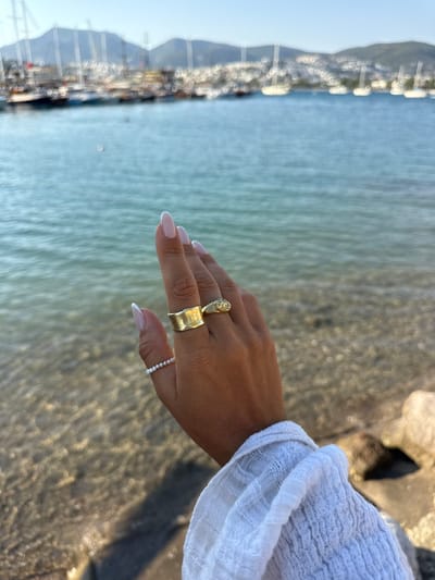 This media file features a close-up shot of a person's hand adorned with rings, positioned in front of a body of water. The hand has well-manicured nails with a light pink polish and a French manicure tip. On the ring finger, there are two rings: a wider, textured gold band and a delicate band with small pearls. Another ring, appearing to be gold with a stone, is worn on the index finger. The person is wearing a white, gauzy garment on their arm, likely a sleeve or shawl.
The background consists of calm, rippling water that reflects sunlight, creating a shimmering effect. To the left, a blurred landscape suggests a shoreline with structures, possibly boats or buildings along a harbor, hinting at a coastal setting. The overall lighting and reflections suggest it is daytime, with bright sunlight.
The scene evokes a sense of relaxation and perhaps a vacation or leisurely activity by the water. The focus on the hand and jewelry, combined with the serene water background, creates an aesthetically pleasing image. The location context of Bodrum, Turkey, reinforces the impression of a seaside destination. No text is visible in the image.