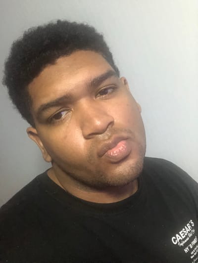 This image captures a close-up portrait of a single adult male, approximately 20-35 years of age, with dark skin and short, dark, curly hair. He is positioned centrally, directly facing the camera with his head tilted slightly to the viewer's left. His expression is characterized by a slight pout, with his lips pursed. His eyes are dark, and a prominent reflection, possibly from a light source or screen, is visible in his left eye (viewer's right). He exhibits stubble on his chin and upper lip. The individual is wearing a black, crew-neck t-shirt. Text is visible on the lower right portion of the shirt, featuring "CAESAR'S" in large letters, followed by "Infamous Hot Dogs," "NY'S FINEST," and what appears to be an address, "148 W 33rd St." The background is a plain, undecorated light grey wall. The scene is illuminated by soft, frontal lighting. This scene is located in the city of Rímac, Peru.