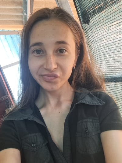 A young woman with long, brown hair and fair skin with freckles smiles for a selfie. She is wearing a dark, button-up shirt. Her hair is parted down the middle and falls loosely around her face. She is looking directly at the camera with a slight smirk. The background is somewhat blurred, but appears to be an outdoor setting with some sort of netting or shade cloth in the foreground. The lighting suggests it is daytime, and the overall mood is casual and relaxed. The image is rotated 90 degrees clockwise.