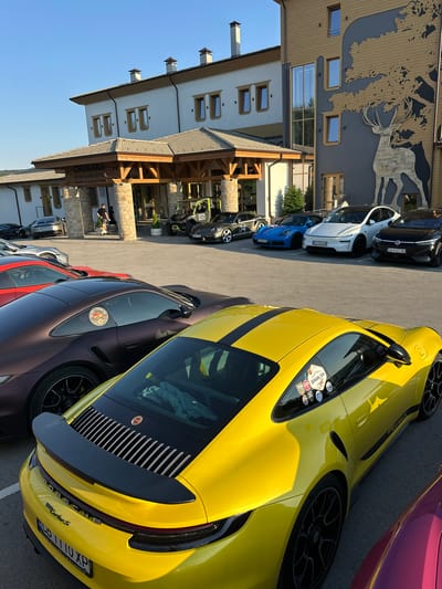 The image shows a row of luxury cars parked in front of a hotel called CHUKARA WINE & SPA HOTEL, located in Kerenite, Bulgaria. The hotel features a modern architectural style with stone and wood accents, and a large graphic of a deer on its facade. The cars visible include several Porsches, a white Tesla, and a black Volkswagen ID.4. One person is walking out of the hotel entrance. The scene is set outdoors under a clear blue sky with the sun casting shadows.