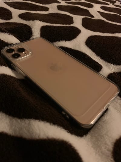 A rose gold iPhone 11 Pro in a clear case lies diagonally on a brown and white cow-print blanket. The phone is positioned with the rear camera array towards the top left corner of the image. The Apple logo is faintly visible on the back of the phone. The blanket has large, irregular dark brown patches on a white background. The lighting is dim, casting shadows. The image was taken indoors, likely in a domestic setting. There is no information available to determine the specific location within Чыжэвіцкі сельскі Савет, Belarus.