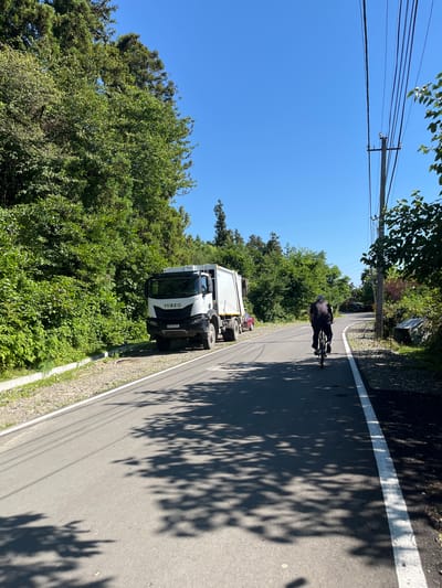 The image depicts a roadside scene on a sunny day, likely in Ureki, Georgia. The main subjects are a white Iveco garbage truck parked on the right side of a paved road, and a person riding a bicycle further down the road. The road has a white line marking its edge. Lush green trees and foliage line the left side of the road, with power lines visible against a clear blue sky. Shadows of tree leaves are cast across the road, indicating it is daytime. The overall impression is that of a quiet, rural or suburban environment during fair weather. The garbage truck has the license plate "AT 972 XX".