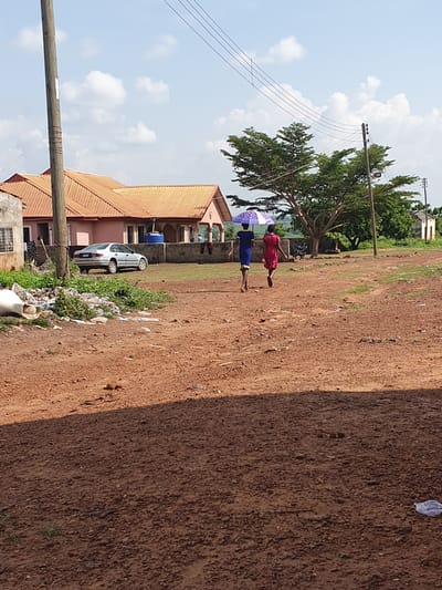 The image, rotated 90 degrees clockwise, captures an outdoor scene in what appears to be a semi-urban or rural area of Idah, Nigeria, under bright daylight.
The foreground is dominated by a wide, unpaved reddish-brown dirt path or ground, showing textures and scattered debris including plastic waste. In the midground, two individuals are walking along this path. One person, dressed in a red outfit, walks slightly ahead of another person wearing a blue outfit. The person in blue holds a purple and white umbrella open, suggesting protection from sun or rain.
To the left of the path, several buildings with distinctive reddish-brown corrugated metal roofs are visible. A white car is parked near one of these buildings. A large tree with dense green foliage stands behind the walking individuals and buildings, casting some shade.
Utility poles with multiple power lines cut across the upper left portion of the frame, set against a bright blue sky dotted with fluffy white clouds. The overall impression is a sunny day in an area with a mix of natural elements, residential structures, and some signs of litter. No discernible text is visible within the image.