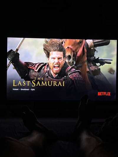 A television screen displays the movie poster for "The Last Samurai." The poster features a close-up of a man with a fierce expression, his mouth open in a shout, holding a sword aloft. He is dressed in samurai armor. Behind him, a horse and other figures are visible, suggesting a battle scene. The Netflix logo is visible in the bottom right corner. The screen is viewed from a low angle, with the legs of a person visible in the foreground, suggesting they are lying down and watching the screen. The overall lighting suggests the room is dark. The location is identified as Mollet Del Vallès, Spain.