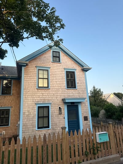 A tan cedar shingled house with light blue trim is the main subject of this image, located at 15 Commercial Street in Provincetown, Massachusetts, USA. The house has multiple windows with dark frames and shutters, and a prominent teal-colored door. A wooden picket fence runs along the front of the property, and a teal mailbox with the number "15" is attached to the fence. Trees and clear blue sky are visible in the background.