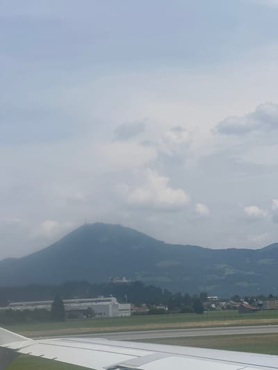 The image shows a hazy aerial view of a mountainous landscape near Salzburg, Austria. A portion of an airplane wing is visible in the foreground, suggesting the photo was taken from an aircraft. The sky is overcast with scattered clouds, creating a soft, diffused light that suggests it is daytime, possibly morning or late afternoon.

In the mid-ground, a dark, imposing mountain range dominates the scene, its slopes covered in dense, dark green foliage. Nestled at the base of the mountains is a small village or town, with scattered buildings, some with red roofs, and patches of green fields. Further down, closer to the foreground, are some larger, modern buildings with white facades and multiple windows, possibly industrial or commercial structures.

Adjacent to the buildings and extending towards the right edge of the image is a striped surface, likely an airport runway. The overall atmosphere is serene and somewhat muted due to the haze and overcast conditions, evoking a sense of peaceful travel. No people are visible, and no text is discernible in the image.