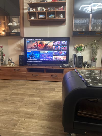 This is a media file showing a living room scene, likely in a home in Villeurbanne, France, oriented vertically. The main focus is a television displaying a video game interface, specifically for GTA Online. The screen shows game promotions and information, including "2X НАГРАДЫ" (2X REWARDS), "БОНУСЫ ЗА ПРОДАЖУ КИСЛОТЫ" (BONUSES FOR ACID SALES), and "СКИДКА В 2 000 000 GTA $" (DISCOUNT OF 2,000,000 GTA$). Other text includes "СРАЗУ К ДЕЛУ" (STRAIGHT TO BUSINESS), "ОБНОВЛЕНИЯ И ОГРАБЛЕНИЯ" (UPDATES AND HEISTS), and "«КОЛЛЕКЦИОНЕР ИСКУССТВА»" (ART COLLECTOR).

In front of the TV, a black speaker and what appears to be a gaming console controller are visible. To the left of the TV is a wooden shelving unit filled with decorative items, including framed photos and figurines. Above this, a dark cabinet with glass doors displays glassware. To the right of the TV, a black, curved piece of furniture is partially visible. The overall setting is a domestic indoor space, suggesting a leisure activity. The lighting suggests it could be daytime, but the artificial lights of the room are also active. There are no people clearly visible in the image.
