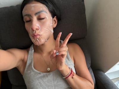 A young woman with a sheet mask on her face is lying back in a gray armchair. She is wearing a gray tank top and a gold necklace with a circular pendant. Her eyes are closed and her lips are pursed. She is holding up two fingers in a peace sign. Her left arm is bent and her hand is near her face. She has a red and clear beaded bracelet on her left wrist. The background is a plain wall and the armchair. The image was taken in Imatra, Finland.