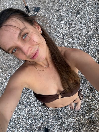 The media file shows a woman lying on a pebble beach. She is wearing a brown bikini and has long, dark brown hair. Her eyes are green and she is looking at the camera with a slight smile. A thin silver necklace is visible around her neck. Her left arm is resting on the pebbles, and a small black object, possibly a shovel, is partially visible near her head. Her toes are also visible, with white nail polish on them. The scene is outdoors, with a clear view of the textured pebble beach. The lighting suggests it is daytime, possibly sunny given the shadows and the overall brightness. The location is identified as Drobnići, Montenegro. The woman appears relaxed and happy. There is no visible text in the media file.