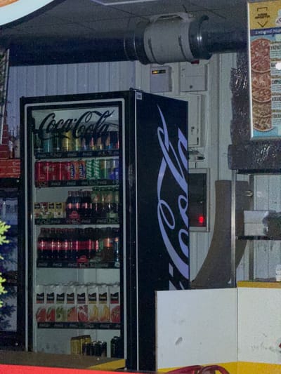 The image shows a refrigerated display case filled with various beverages, primarily Coca-Cola products, situated in an outdoor setting. The display case is black with a large white Coca-Cola logo on the left side. Inside, rows of Coca-Cola cans and bottles are visible, along with some Fanta and Sprite. On a shelf below, there are juice boxes labeled "Cappy". The background suggests an urban environment, possibly a street or an outdoor market area, with a corrugated metal wall and a sign with images of pizzas on the right. The lighting indicates it is likely evening or nighttime. The overall scene appears to be a point of sale for beverages. There are no people visible in the frame. The location is identified as Stryi, Ukraine.