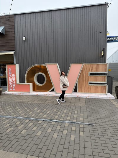 An outdoor, eye-level photograph depicts an adult female posing in front of a large "LOVE" sign at Hede Fashion Outlet in Kungsbacka, Sweden. The woman stands slightly right of center, facing partially towards the camera with a smile, her arms crossed, hands tucked into the sleeves of her cream-colored, mid-thigh-length quilted jacket. She wears black trousers and athletic shoes with white, black, and gold accents, and carries a small brown shoulder bag.
The oversized "LOVE" sign is composed of four distinct letters, each approximately human-height. The "L" is a coral-pink box displaying "FOREVER PIECES" in white vertical text. The "O" is a reflective gold/bronze material. The "V" is coral-pink, and the "E" is a light wood-grain finish. All letters feature small, illuminated points along their edges and rest on a white base speckled with pink details.
The background consists of a tall, dark grey building with vertical siding. To the left, a section of this building features brown horizontal siding and an awning over a glass entrance. To the right, a grey corrugated building is partially visible, alongside a dark planter and a yellow warning sign on a pole. The foreground is paved with rectangular grey and lighter grey blocks arranged in an alternating pattern, bisected by a linear drainage grate. An overcast sky is visible, with a string of small lights suspended overhead.