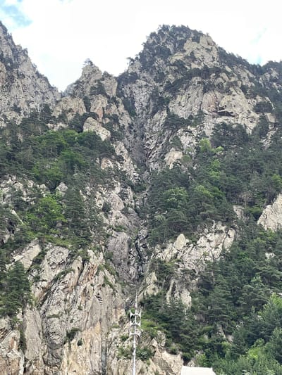 This is a close-up, ground-level, eye-level shot of a rugged, rocky mountain face covered in dense pine trees. The mountainside is steep and uneven, with large outcroppings of grey rock interspersed with patches of greenery. The trees are a mix of dark green and lighter green, indicating different species or ages. In the lower right of the frame, a white, multi-pronged antenna-like structure rises vertically, extending from the base of the mountain. The sky above is overcast with patches of white cloud. The overall impression is one of wild, untamed nature, with the artificial structure of the antenna providing a stark contrast. This scene is consistent with the landscape of Dariali Gorge, Georgia, which is known for its dramatic mountain scenery and the presence of infrastructure like communication towers.