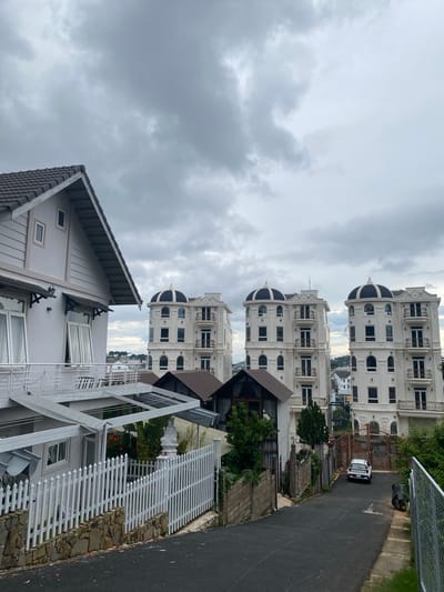 The image depicts a street scene in the Lam Vien area of Da Lat, Vietnam, under a cloudy sky. On the left, a light-colored, multi-story house with a pitched roof is partially visible, featuring large windows and a balcony. In the center and right of the image, three identical white buildings with domed roofs and ornate balconies stand in a row. A white car is parked on the asphalt road in front of a large, wrought-iron gate. The road is lined with fences and vegetation.