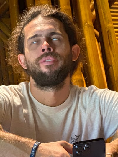 The image, set in Mù Cang Chải, Vietnam, captures a male individual from the chest up, positioned against a backdrop of vertical yellowish-orange bamboo poles. He possesses dark, curly, shoulder-length hair and a full, dark beard and mustache. His facial expression is distinctive, with his eyes rolled upwards, revealing primarily the whites (sclera) and a sliver of blue iris. His mouth is slightly open, with the upper lip slightly raised, showing his upper teeth.

He wears a light beige, short-sleeved t-shirt with a subtle texture, featuring a small, dark, stylized animal logo (resembling the Arc'teryx emblem) on the left chest area. His forearms are visible, appearing tan and hairy. On his left wrist, he wears a blue fabric bracelet with repeating white diamond-like patterns. His right hand holds a dark-colored smartphone, seen from the rear, prominently displaying a multi-lens camera module. The lighting is strong and directional, illuminating the man's forehead and casting shadows under his eyes and chin. The bamboo poles in the background are arranged vertically, creating a textured, natural wall or screen.