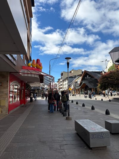 A slightly angled view of a pedestrian street in San Carlos de Bariloche, Argentina, with a bright blue sky and scattered clouds above. The street is paved with gray rectangular stones. On the right side, a row of dark, rounded bollards separates the pedestrian area from the street. Concrete benches with graffiti are also visible. Several people are walking along the street, dressed in casual clothing, some in jackets and jeans.

On the left, the corner of a building is visible, with a red-framed storefront adorned with colorful balloons. A store sign with some lettering can be seen. Further down the street, buildings with dark, pitched roofs can be seen, including "EL REINO de los Chocolate". Streetlights are present along the thoroughfare, and wires extend across the sky.