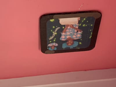 A framed photograph is displayed on a pink wall, likely in Kakau, Nigeria. The photograph features a young woman in traditional attire, including a blue headscarf and a patterned blue and pink dress. She is standing in front of a cake, with her hands positioned as if to cut it. The background of the photo is dark with scattered green and yellow confetti-like shapes. Above the main image, a smaller inset photo shows a close-up of the same woman's face, also wearing a blue headscarf. White text, curving across the photograph, reads "HAPPY BIRTHDAY." The overall impression is a celebratory and personal moment captured in time. The lighting in the original image suggests indoor illumination. The pink wall and the somewhat worn frame of the photograph suggest a lived-in domestic setting.