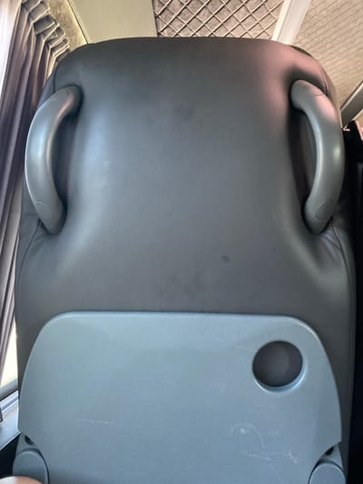 The media file shows a close-up, angled view of the interior of a vehicle, possibly a bus or van, with a focus on a gray, possibly leather, seat. The seat has two prominent gray handles, one on the left and one on the right side, which appear to be part of the seat's structure or for passenger use. Attached to the right side of the seat is a fold-out tray table, also in a gray color, with a circular indentation or holder. The background shows a quilted gray material, likely upholstery on another part of the vehicle's interior, and pleated gray fabric at the bottom, possibly curtains or seat coverings. The lighting suggests daytime. There is no visible text in the image. The scene appears to be static, with no activity taking place other than the presentation of the seating area. The overall impression is of a functional vehicle interior, likely in a public transportation context. The location is identified as Żółtki, Poland.