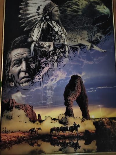 This media file is a framed artwork depicting a composite scene. At the top, a Native American chief in full headdress is visible, alongside a large eagle and a wolf. Below them, a superimposed portrait of an elder Native American man's face emerges from swirling clouds. The background features a dramatic sky with a large rock arch formation, reminiscent of natural arches found in arid desert landscapes. In the foreground, a serene body of water reflects the sky and the rock arch. Two horses with riders are depicted in the middle ground, traversing a rocky, arid terrain. The overall scene evokes a sense of the American West, with its iconic imagery of Native American culture, wildlife, and dramatic natural landscapes. The time of day appears to be either dawn or dusk, with a warm, golden light illuminating the horizon and casting reflections on the water. The atmosphere is somewhat dramatic, with stormy clouds in the upper sky contrasting with the calmer, reflective foreground. No visible text is present in the artwork.