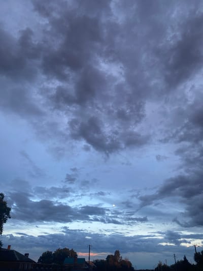 The image shows a dramatic sky filled with dark, heavy clouds. The clouds are varied in texture and density, with some areas appearing almost black while others are lighter shades of gray. A faint crescent moon is visible in the sky, appearing as a small, pale disc. The overall impression is one of an approaching storm or the aftermath of one.

The scene is likely outdoors, as suggested by the presence of trees and power lines on the right side of the frame, along with the suggestion of rooftops of houses. The time of day appears to be either dusk or dawn, given the subdued lighting and the presence of the moon. The weather is overcast and possibly stormy.

The image is taken from a low angle, looking up at the sky. The composition emphasizes the vastness and power of the clouds. There is no visible text in the image. The emotion evoked is one of awe and perhaps a touch of foreboding due to the dark clouds. The location context provided is Yegorlykskaya, Russia, which aligns with the rural or semi-rural feel suggested by the scene.