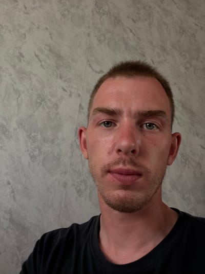 A close-up, eye-level, waist-up photograph shows a young man from Mishkino, Russia. He has short brown hair and is looking directly at the camera. He has light skin with some redness on his cheeks, and a light stubble beard. He is wearing a black t-shirt. The background is a gray, textured wall.