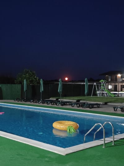 This is a nighttime outdoor shot of a swimming pool area. The pool is illuminated from within and is a vibrant blue. A yellow inflatable ring, shaped like a pineapple, floats in the water, with a green float partially submerged. Pool ladders are visible at the edge of the pool. The surrounding area is covered in green turf. Behind the pool, a row of closed green umbrellas stands next to several empty lounge chairs. A fence runs along the back of the lounge area, with trees visible beyond it. Further back, a playground with a slide and swings is present, and a building with lit windows can be seen in the distance. The sky is a deep indigo, with a bright, full moon visible in the upper left portion of the frame. The overall impression is one of a serene and tranquil evening setting, likely at a resort or recreational facility in Shkurupiivka, Ukraine. There is no visible text.