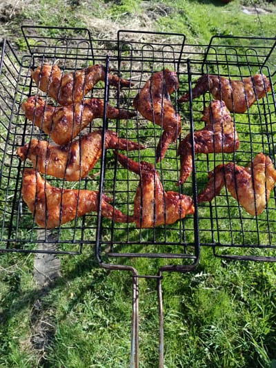 The image shows a grill basket filled with cooked chicken wings. The grill basket is sitting on grass, suggesting an outdoor cooking setting. The wings appear to be well-cooked, with a golden-brown color and some char marks. The grill is positioned on top of a green grass field. The location given is Вичелово, Russia, but there are no specific landmarks or indicators within the image to confirm the city.