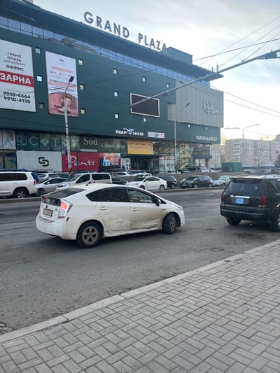 This media file captures a street scene in Ulaanbaatar, Mongolia, featuring a multi-story commercial building and a line of parked cars. The image is taken from a slightly elevated perspective, looking down the street.
The prominent building on the left side of the frame is dark gray or dark green-gray with numerous windows and has "GRAND PLAZA" in large English letters on its upper facade. Various commercial signs adorn the building, including "SUPERMARKET," "МИРНОМ" (MIRNOM), "CINOPING," "Classic shop," "Sod," "Penti," and "CLINIQUE." A large banner advertises "ГРАНД ПЛАЗА ЦОГЦОЛБОРТ ОФФИС ЗАРНА" (GRAND PLAZA COMPLEX OFFICE FOR SALE) with phone numbers "9910-4664" and "9910-4130," and another mentions "СУПЕР СУГАЛААТАЙ" (SUPER LOTTERY/RAFFLE).
Along the street in front of the building, numerous cars are parked. A white Toyota Prius is prominently featured in the foreground, facing away from the viewer. Behind it, other cars, including a black SUV with a license plate partially visible as "4958," line the street. The vehicles appear to be parked parallel to the curb.
To the right, a paved sidewalk with rectangular pavers laid in a herringbone pattern runs parallel to the street. Overhead power lines and utility poles are visible against a bright, slightly hazy sky, suggesting daylight hours, possibly late morning or early afternoon. No specific activity involving people is clearly discernible, but the urban setting with commercial establishments implies a regular day of business.