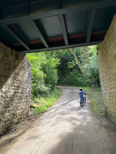 A person on a bicycle rides along a paved path that runs through a tunnel or underpass in Pétange, Luxembourg. The left side of the image is dominated by a dark green, steel structure, likely part of the bridge or tunnel's support system. To the right, a weathered brick wall forms the side of the tunnel. Beyond the tunnel's opening, a densely vegetated area is visible, with lush green trees and plants lining the path. The cyclist is wearing a helmet and a blue jacket. The path is paved, and dappled with light and shadow. The overall scene suggests a natural or park-like environment, characteristic of Pétange, Luxembourg, where such green spaces often intersect with infrastructure.