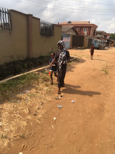 A daytime scene depicts an unpaved, reddish-brown dirt road in Egan, Nigeria, under an overcast sky. Three individuals are visible. In the foreground, an adult female wearing a patterned headscarf and a dark, patterned long dress walks towards the viewer's right, casting a prominent shadow. She wears sandals and holds an object in her left hand. A young child, wearing a dark blue shirt and light shorts, walks slightly behind and to the left of the adult, holding a light-colored object. An orange traffic cone is present on the ground near this child. To the left of the road, a low concrete curb borders a strip of green grass and dry vegetation. Beyond this, a tall, tan concrete wall with dark metal spiked fencing on top runs parallel to the road. Scattered litter, including plastic wrappers, is visible on the dirt road and in the vegetation. In the mid-ground, a second child, dressed in reddish-brown attire, walks further down the road on the right side. A light blue van or small bus is parked or stationary further down the road. Various residential buildings, including a two-story structure with a reddish-brown roof, are visible in the background. Overhead power lines traverse the sky.