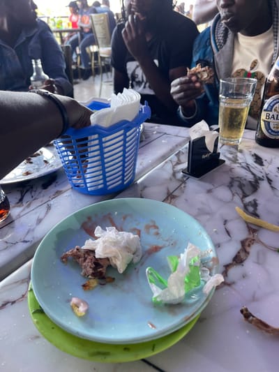The image captures a casual dining scene, likely in an indoor or semi-outdoor restaurant setting, featuring several individuals gathered around a marble-patterned table. The perspective is an overhead, slightly tilted view, showing the table surface and the upper bodies of some patrons.
In the upper left, a person with dark skin wears a light-colored top with a patterned design, holding food in their hands. Next to them is a glass containing a light-amber beverage and a brown bottle with "BALTICA LAGER BEER" clearly visible on its label. Another person, wearing a black shirt, is seated further down on the left, also engaged in eating.
The foreground on the right features a person's arm and hand holding a blue plastic basket filled with white paper napkins. To its right, a light blue plate is stacked on a green plate. The light blue plate is mostly empty, showing remnants of food, sauce smears, and crumpled white and light green used napkins. A small portion of meat or stew residue is visible on the plate.
On the table surface, between the diners and the plates, there is a small black holder, possibly for napkins or receipts, with some faint white text that is not legible. In the background, other people are visible, along with glimpses of tables and chairs, suggesting a busy environment. The lighting suggests it is daytime. The overall atmosphere appears relaxed, indicative of a social gathering or a meal in a local eatery in Kimorori, Kenya.