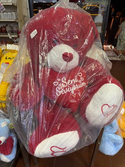 In Acıpayam, Turkey, a large, red teddy bear is displayed prominently, sealed in clear plastic packaging. The bear has white paws and a white muzzle, with red detailing on its snout. Text embroidered in white on its chest reads "Seni seviyorum," meaning "I love you" in Turkish. A price tag with "3000 TL" is attached to the packaging near the bear's head. The teddy bear is positioned on a display rack. To the left, a smaller blue teddy bear also in plastic packaging is visible. In the background, shelves stocked with various items and a shop interior with customers can be seen. The overall scene suggests a retail environment.