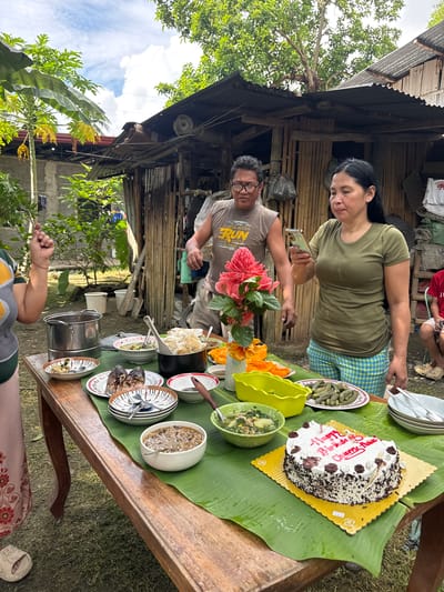 An outdoor birthday celebration is underway in Carmen, Philippines, centered around a long, rustic wooden table covered with banana leaves. A white and chocolate birthday cake, inscribed with "Happy Birthday Chery Anne" in red icing, is prominently displayed on a gold cake board at the table's front right. Multiple dishes are arranged on the table, including a large black pot of white, lumpy food, bowls of green vegetable soup, a brown stew, a plate holding two grilled fish, a plate of small green elongated items, and sliced orange fruit. Various ceramic plates, bowls, and cutlery are set for dining, and a vibrant bouquet of pink torch ginger flowers serves as a centerpiece.

Three adults are clearly visible, with a fourth individual partially present on the far right. On the left, a person wearing a multi-colored (green, yellow, grey) top and a light pink skirt raises their left hand in a gesture. In the center-left, a man with glasses, wearing a grey t-shirt, stands behind the table, looking towards the right. In the center-right, a woman with long dark hair, dressed in an olive green t-shirt and blue and yellow plaid shorts, stands behind the table, engrossed in a smartphone held in her right hand. A partially visible individual wearing a red shirt and light-colored clogs is seated on the far right.

The environment consists of grassy ground and abundant tropical vegetation, including a papaya tree with yellowing leaves and fruit on the left. In the background, a rustic structure constructed with bamboo walls and a corrugated metal roof is visible, alongside a concrete block wall building where clothes are hanging to dry. The sky above is partly cloudy blue, indicating daylight.