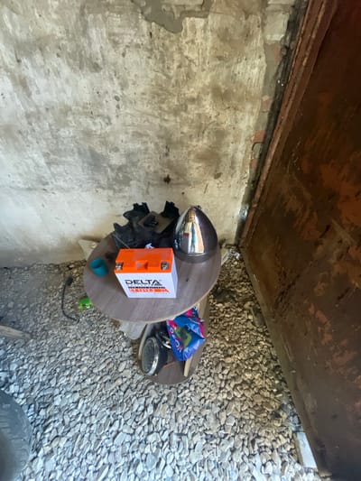 An overhead shot shows a motorcycle battery, a chrome-plated helmet, and other miscellaneous items placed on a small, round table situated on a bed of gravel. The table, constructed from wood, has a visible grain pattern and appears to be set against a rough, concrete wall. The wall exhibits signs of wear and tear, with patches of exposed brick and crumbling plaster. To the right of the scene, a large, rusty metal door or panel dominates the frame. The lighting suggests an indoor environment, possibly a garage or storage area, with the sunlight creating shadows. The overall impression is one of a somewhat neglected or utilitarian space.