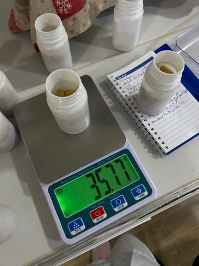 The image depicts an overhead view of an indoor scene, presumably in Oran, Algeria, where a digital scale is being used to weigh items. A Ming Heng Digital Scale (Model: MH-885) displays a reading of "35.77 g" with a precision of "d=0.01g" and a maximum capacity of "Max=600g". Positioned on the scale's silver platform is an open, white, cylindrical plastic bottle containing multiple yellow, elongated capsules.
To the right of the scale, an open, spiral-bound notebook with ruled pages is visible. Handwritten entries include "Note Book", "Screaufou", "Royal 60 Oel", and various numerical values such as "34,63", "34,27", "30,80g", "36,42", "30,30", and "36,77". Another identical open white plastic bottle, containing a single yellow capsule, rests beside the notebook. Additional empty white plastic bottles are scattered in the upper background.
A red and white patterned cloth, possibly featuring snowflake designs, is partially visible in the upper left background. A clear plastic container or lid is situated in the upper right. The underlying surface is a light-colored countertop or table. In the lower right foreground, partial views of a person's legs and feet are visible, with one foot wearing a shoe that has a pink sole.