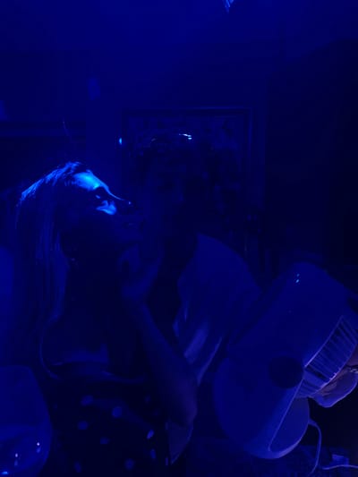 This image depicts two individuals in what appears to be an indoor setting, possibly a party or gathering, illuminated by a strong blue light. The focus is on the close interaction between a woman in the foreground and a person behind her. The woman, with her head tilted upwards and eyes closed, has her face partially illuminated by the blue light, highlighting her features. She is wearing a dark garment with a polka dot pattern. The person behind her is silhouetted and their features are not clearly discernible, but they appear to be leaning towards the woman.
In the lower right portion of the image, a white fan is prominently featured, its blades and casing visible under the blue light. The background is dark and indistinct, with hints of other objects or decorations, but nothing is clearly identifiable. The overall atmosphere is intimate and perhaps slightly mysterious due to the low lighting conditions and the intense blue hue.
The location provided, Pozuelo de Alarcón, Spain, does not offer any specific contextual clues visible within the image itself. The scene is generic and could be anywhere a gathering with blue lighting and a fan might occur.