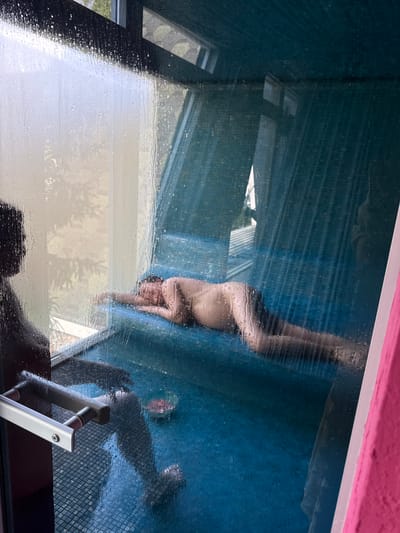 The image shows a blurred reflection of a person in a window. The person appears to be lying on a blue surface. Outside the window, there are glimpses of buildings and greenery. The window has a handle. The upper frame of the window is painted pink. The image location is Чёрное, Russia.