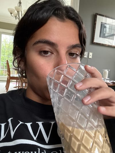 A close-up portrait captures a young adult female from the chest up, positioned indoors. She has dark hair and dark eyes, looking directly at the viewer. Her face is partially obscured by a clear, diamond-patterned drinking glass held in her right hand, from which she appears to be consuming a light brown liquid. She wears a black crew-neck t-shirt with partial white lettering, "WW", visible on the chest.
The background depicts a residential interior. To the left, a large window allows natural light, revealing green foliage outdoors, along with wooden dining chairs and a table. A multi-arm chandelier with light-colored shades hangs from the ceiling. To the right, a dark grey wall displays a framed picture, an electrical wall outlet, and a white cylindrical object, possibly a candle, on a flat surface.
There are no discernible visual cues within the image to establish the specific location as the city of Princeton, United States; the scene presents a generic indoor residential environment.