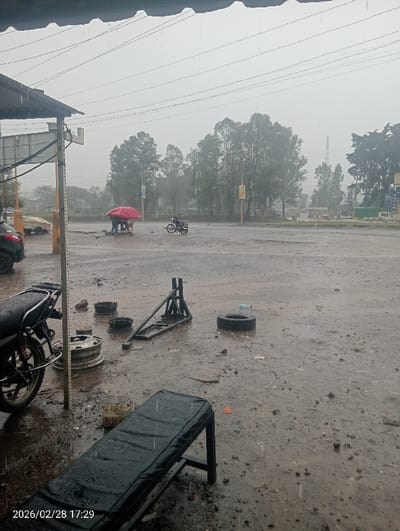 The scene depicts an outdoor area in Kapkures ward, Kenya, captured at 17:29 on 2026/02/28, during a rainfall event. The ground consists of wet, muddy earth and standing water, reflecting the overcast sky.
In the immediate foreground, a black, padded bench is visible, covered in raindrops. To its left, the rear wheel and partial frame of a black motorcycle are present. Further into the mid-ground, three bare metal wheel rims and a black metal frame, possibly a vehicle stand or part, lie on the wet ground. A single car tire rests flat near a clear plastic bottle.
In the center-midground, two individuals are observed sheltering under a large red umbrella. One appears seated, the other standing. To their right, a black motorcycle equipped with a cargo attachment is stationary on the wet, unpaved surface. Beyond this area, a paved road or larger open area extends.
The background features numerous trees, appearing somewhat indistinct due to the rain and distance. Utility poles with overhead power lines are visible across the sky. Several vehicles, including a light-colored car, are partially visible in the distance on the left. A tall, slender structure is faintly discernible on the right horizon, partially obscured by trees. The entire environment is saturated with rain, creating a hazy atmosphere.
