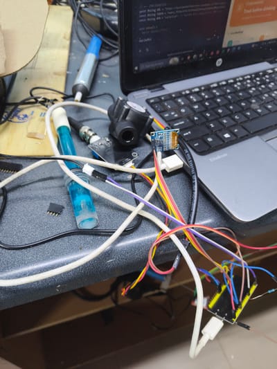 The image shows a cluttered workspace on a dark gray countertop. Dominating the scene is an open HP EliteBook laptop with a webpage displayed showing code with the URL "https://you". A white USB cable connects the laptop to a small, blue circuit board. Various wires, predominantly white, yellow, red, and blue, connect this circuit board to a second, larger black circuit board located at the lower right of the image.
Other items visible include a pen with a blue cap, a pH sensor with a clear body and blue liquid, a black sensor, and various electronic components scattered across the surface.
There is nothing in the image to suggest the location is in Thika Township Ward, Kenya.