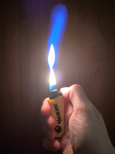 A person's hand holds a yellow lighter with a blue flame. The lighter has black accents and text that reads "ЧИЖИК" in black Cyrillic letters, along with a small depiction of a bird. The background is dark and indistinct. The image does not contain any specific location information for Protvino, Russia.