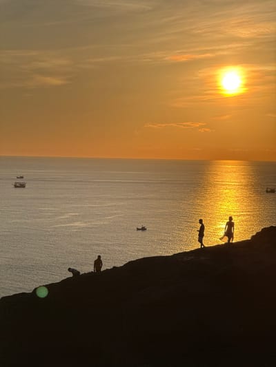 The image depicts a serene sunset over the sea, viewed from a rocky coastline. The sky is a warm gradient of orange and yellow, with wispy clouds scattered across it. The sun, a bright orb, is positioned high in the sky, casting a golden reflection on the rippling water. Several boats are visible on the sea, appearing as dark silhouettes against the bright expanse.
In the foreground, the dark silhouette of a rocky cliff overlooks the ocean. Several figures are visible on the cliff edge. Two individuals stand close together, facing the sea. One appears to be a man with short hair, wearing shorts. The other, possibly a woman, is seen from the back, wearing a flowing dress. Further down the cliff, another figure is bent over, as if looking at something on the ground. A fourth figure, standing, is further to the left, silhouetted against the sea. A faint green light anomaly is visible on the cliff face in the lower left quadrant of the image.
The scene suggests a peaceful moment of observing nature's beauty, likely a popular viewpoint on the island of Lý Sơn, known for its stunning coastal scenery and sunsets.