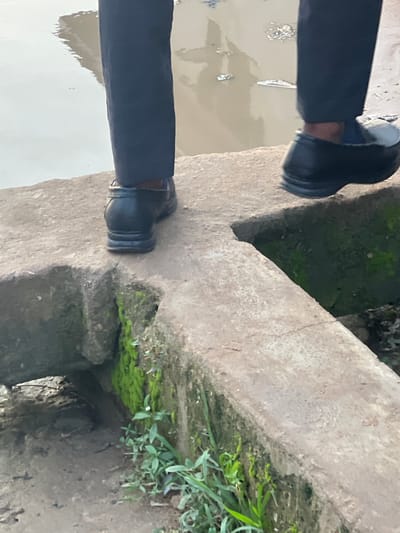 The image presents a ground-level, slightly tilted view of a person's lower legs and feet. The individual is wearing black trousers and black, somewhat chunky formal shoes. They are positioned on a rough, grey concrete structure, which appears to be a bridge, walkway, or part of a drainage system.

The left side of the image shows a body of murky, brownish-grey water, possibly a canal or large puddle, reflecting the light from above. Floating debris is visible on the water's surface. The person's left leg and foot are near the edge of this water body.

The right side of the image reveals more of the concrete structure, which is partially covered in vibrant green moss and small, sparse green plants. There is a distinct gap or opening in the concrete below the person's right foot, leading into a dark, shadowed recess. The overall texture of the concrete is weathered and uneven.

The scene suggests an outdoor environment during daylight hours, possibly in a semi-urban or rural area like Ijegun, Nigeria, where such concrete structures are common over waterways. The person appears to be either standing on or carefully traversing this structure. No text is visible in the image.