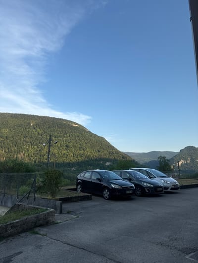 A daytime, eye-level, outdoor shot captures a parking lot in the city of Saint-Claude, France, nestled against a backdrop of lush green mountains. In the foreground, three cars are parked in a row: a black Ford Focus, a dark grey Peugeot 308, and a white Citroën C4 Picasso. The cars are facing to the right of the frame. A chain-link fence runs along the left side of the parking lot, separating it from a concrete structure and some overgrown vegetation. Behind the fence, the mountains are densely covered with trees, their slopes reaching towards a clear blue sky with scattered wispy clouds. A lamppost stands near the center of the frame, between the fence and the cars, with two lights. The ground in the parking lot is asphalt, showing signs of wear and cracks. The overall impression is one of a serene, natural setting adjacent to a developed area.