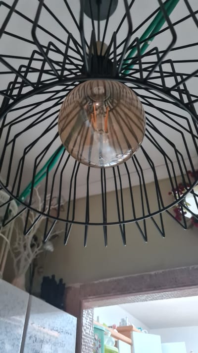 A close-up shot focuses on a vintage-style light fixture with a cage-like black metal frame surrounding an amber-toned light bulb. The bulb’s filament is visible. Green tubing is partially visible, woven through the fixture's metal frame. In the background, a window provides a glimpse of a child's room, complete with a crib, toys, and a whiteboard. The room appears to be in a domestic environment. There is no information about the location being Nahariyya, Israel.