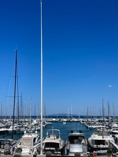 In Punta Ala, Italy, a marina is captured under a clear, bright blue sky. The scene is dominated by a tall, white flagpole in the foreground, casting a slight shadow. To the left, a tall, dark mast of a sailboat stands prominently. Numerous yachts and sailboats are moored in the harbor, filling the frame from the waterline upwards. The boats vary in size and design, with many featuring white hulls and masts. The water is a deep blue, reflecting the sky. In the distance, a hazy landmass can be seen on the horizon. A few small clouds are present in the sky. The bottom portion of the image shows the decks and sterns of several luxury yachts. One yacht prominently displays the name "Chebellea". A few individuals are visible on the docks and on one of the yachts, engaged in casual activity. The overall atmosphere is one of a sunny day at a well-appointed coastal resort.