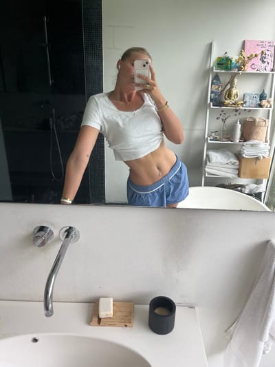 A female subject is captured in a mirror selfie within a bathroom in Skanderborg, Denmark. Her face is obscured by a light pink smartphone held in her left hand, which is actively taking the photograph. She wears a white ribbed short-sleeved crop top, revealing her midriff, and blue shorts with white trim. A navel piercing is visible. A gold watch is on her left wrist, and a white and gold bracelet is on her right wrist. A single white earbud is in her left ear, and a gold ring is on her left ring finger.
The reflected environment includes a dark, possibly tiled, shower area with a black shower fixture to the left. To the right, a white five-tiered ladder shelf displays various items, including a golden Buddha statue, a framed pink text art piece, a small green bottle, dried yellow flowers, a woven basket, and folded white towels. A white bathtub is partially visible in the far right of the reflection. The foreground shows a white countertop with a sink basin. Chrome wall-mounted faucet and controls are present. On the countertop, a wooden soap dish holds a white bar of soap, and a black cylindrical object, possibly a candle holder, is also present. A white towel hangs to the right of the counter.