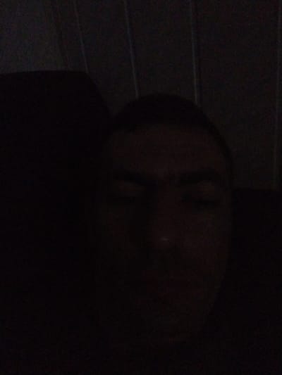The image is a dark, close-up, selfie-style photograph of a person's face. The lighting is very poor, making it difficult to discern many details. The person appears to be looking directly at the camera with their eyes open. There is a faint hint of a headboard or textured background behind them, possibly made of wood or fabric. The scene is likely indoors, given the apparent texture and the close proximity of the background. The specific location in the city of Vylok, Ukraine, cannot be determined from the visual information provided.